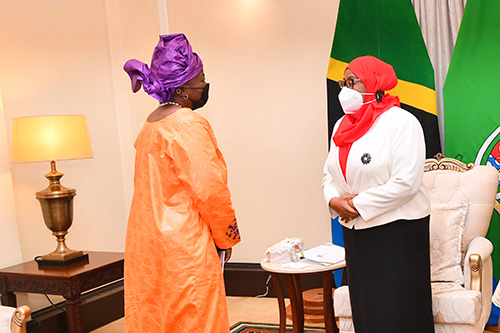 Two women in formal business attire stand apart, wearing face masks. Behind them are the national flag of the United Republic of Tanzania and the flag of the president.