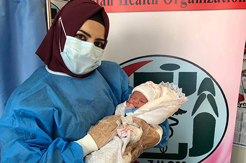 A midwife in a red headscarf and surgical face mask holds onto a newborn baby.
