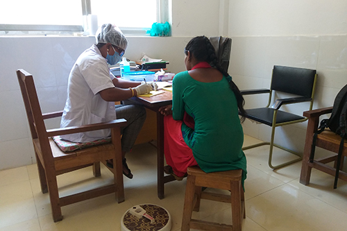 Kala Chaudhary, wearing a face mask, gloves and hair covering, sits at a desk and takes notes while providing a family planning consultation to a woman.