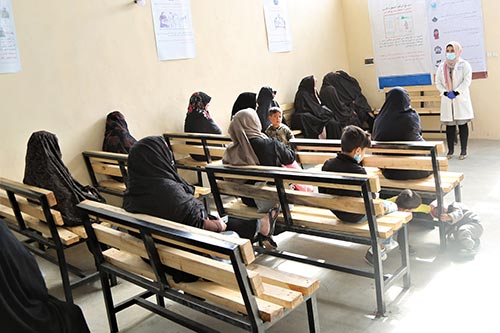 Women in head scarves sit on wooden benches. They are listening to a woman in a white medical coat wearing a face mask.