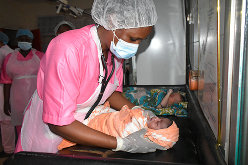 A midwife in Guinea wraps a newborn in a blanket. She is wearing pink scrubs, gloves, a face mask and an apron. 
