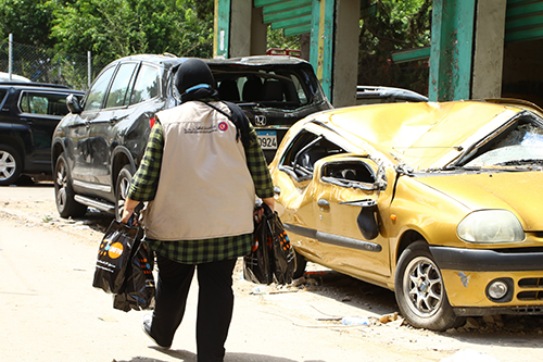 Hiba Kchour carries bags of hygiene supplies past a crushed yellow car.