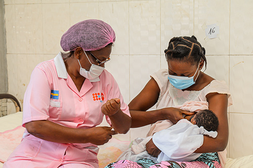 A midwife in a face mask and hair cover helps a new mother learn to breastfeed. The new mother is also wearing a facemask. 