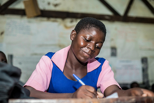 Laureen smiles while writing at a school desk.