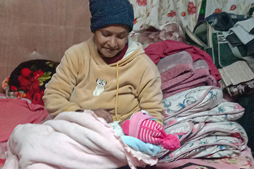 Maya holds her newborn baby girl. Maya is wearing a blue had and her baby is bundled in pink blankets. 