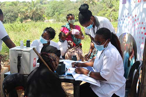 Health workers in face masks speak with patients at an outdoor table. They are part of a mobile clinic. 