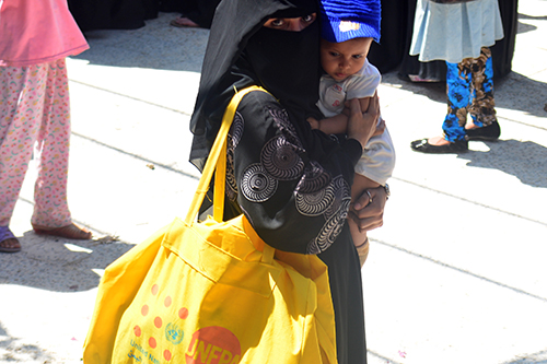 A woman in a niqab holds a baby on one side and carries a yellow dignity kit on the other.