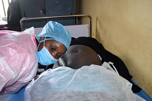 A midwife wearing a mask and hair covering listens to a pregnant woman's abdomen with a pinard stethoscope. 