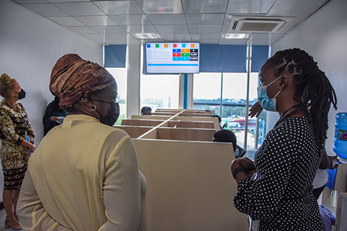 Two women speak in the foreground. Both are wearing face masks. Behind them is a series of cubicles where call centre counsellors are sitting. 