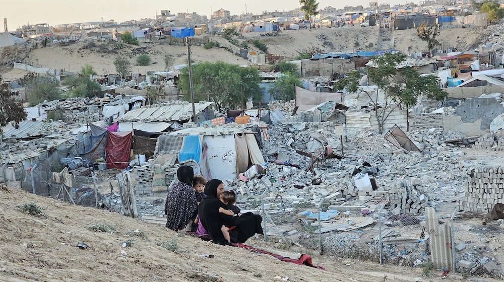 wo women in dark clothing each hold a child while sitting on a hillside overlooking makeshift tents amid the rubble of destroyed buildings.