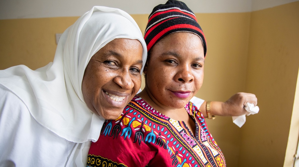 Une femme portant un foulard blanc passe le bras sur les épaules d’une femme en robe rose et jaune vif. Toutes deux sourient.