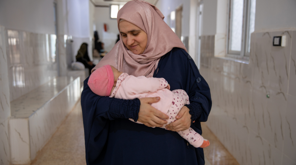  Une femme sourit, tenant un petit bébé dans ses bras, dans le couloir d’un hôpital