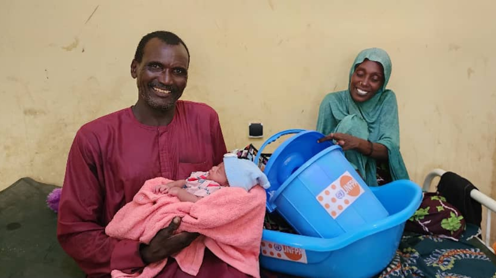 Un homme vêtu de rouge tient un nouveau-né dans ses bras. Derrière lui, la mère de l’enfant, assise sur un lit et vêtue de bleu, tient une petite baignoire bleue avec le logo de l’UNFPA.