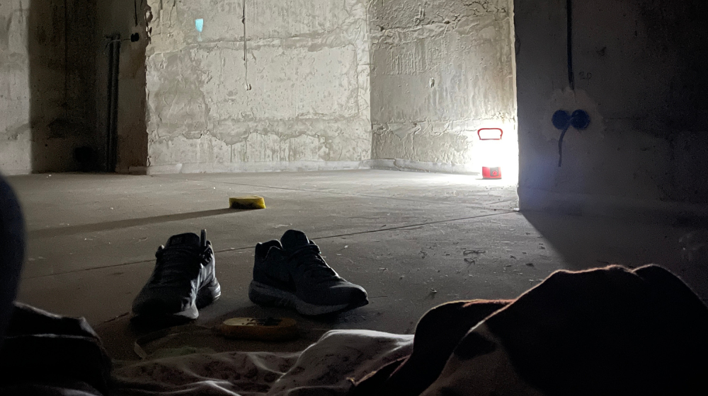 A pair of men’s shoes can be seen in front of some crumpled blankets in an underground bunker. A lamp lights up the corner of the corncrete walls