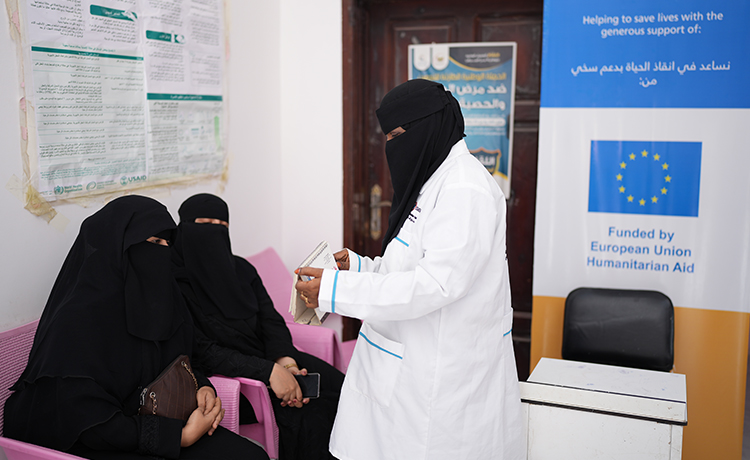 Une sage-femme debout devant deux femmes dans un centre de santé, tenant un bloc-notes.
