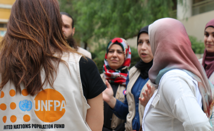 Une femme vêtue d’un gilet portant le logo de l’UNFPA s’entretient avec un groupe de femmes et un homme dans une cour extérieure.