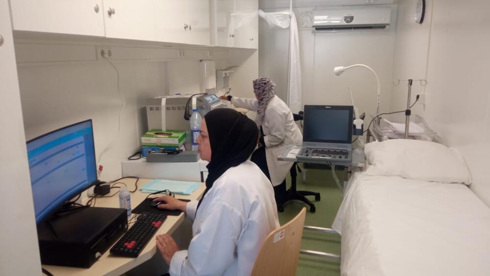 Dos trabajadoras sanitaria sentadas junto a una cama vacía, una mira la pantalla de una computadora, la otra toma una lectura de una máquina