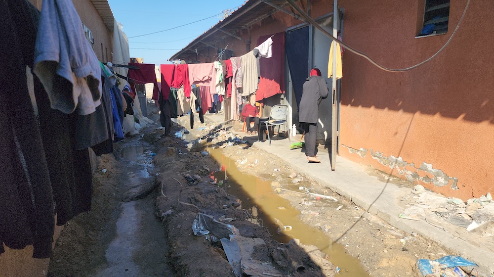 In an alleyway full of dirty water and rubbish, a woman is seen hanging laundry between two houses.