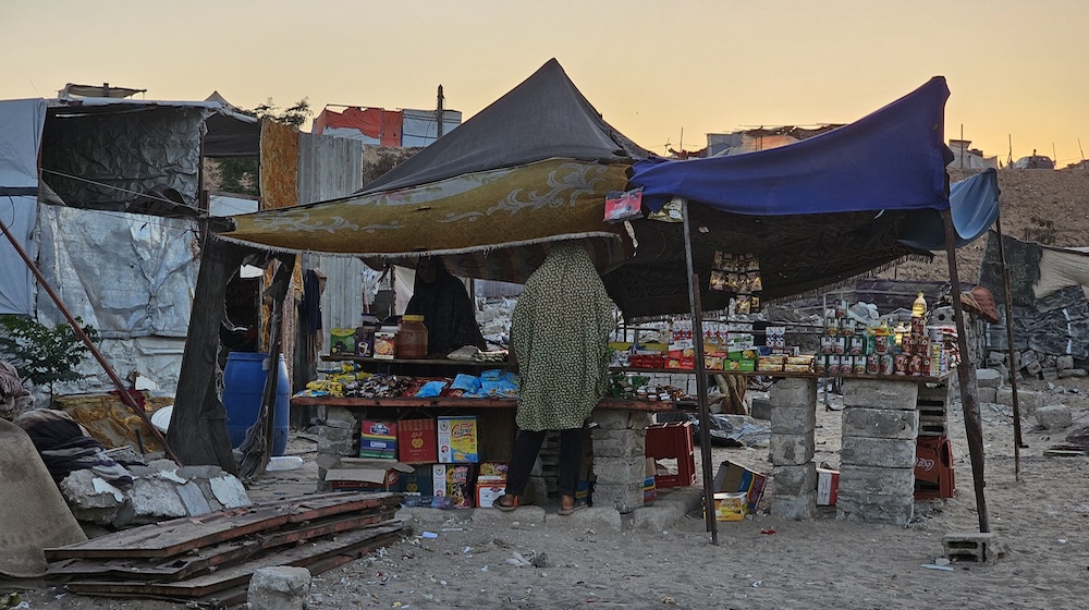 Stocks of packaged food are piled on makeshift tables under tent structures in an informal camp. A woman can be seen from the back wearing a patterned shawl. 