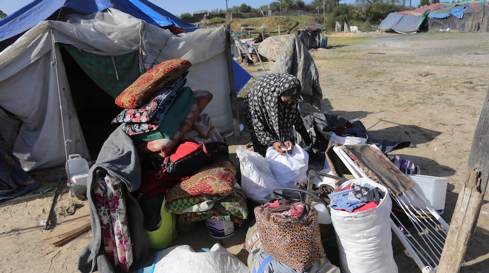 A woman in a patterned head scarf leans over piles of bags and belongings outside a blue and white tent in a dry and dusty field