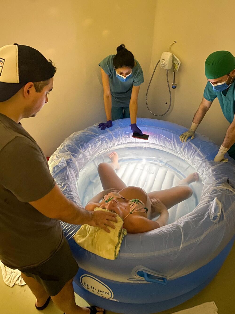 A pregnant woman in a birthing pool with a man outside behind her and two health-care workers in front