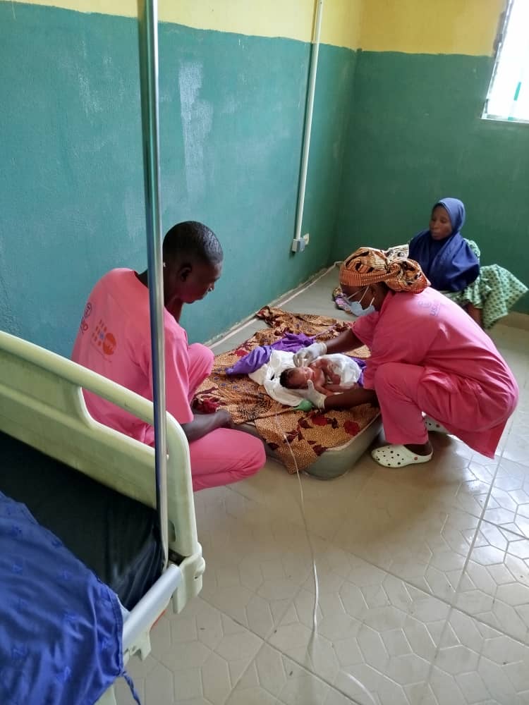  Health-care workers crouch to attend to a newborn baby on a mattress on the floor of a room, with a women seated watching on.