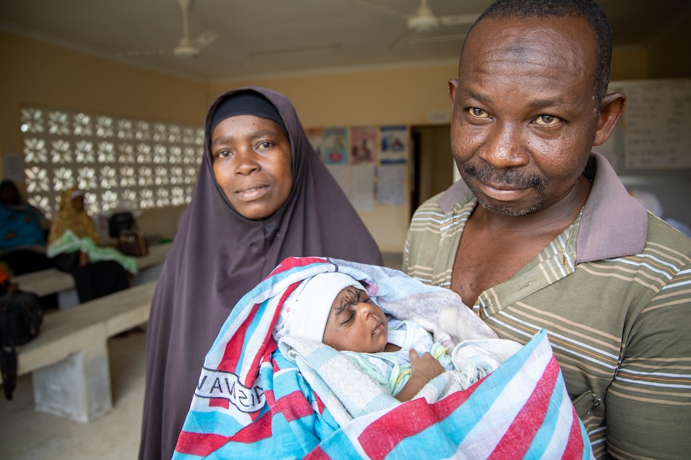 Un homme en polo rayé tient son nouveau-né enveloppé dans une couverture. La mère de l’enfant, vêtue d’un hijab noir, se tient à ses côtés, souriante.