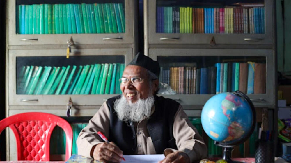 Un homme tient un crayon, assis à un bureau devant une bibliothèque.