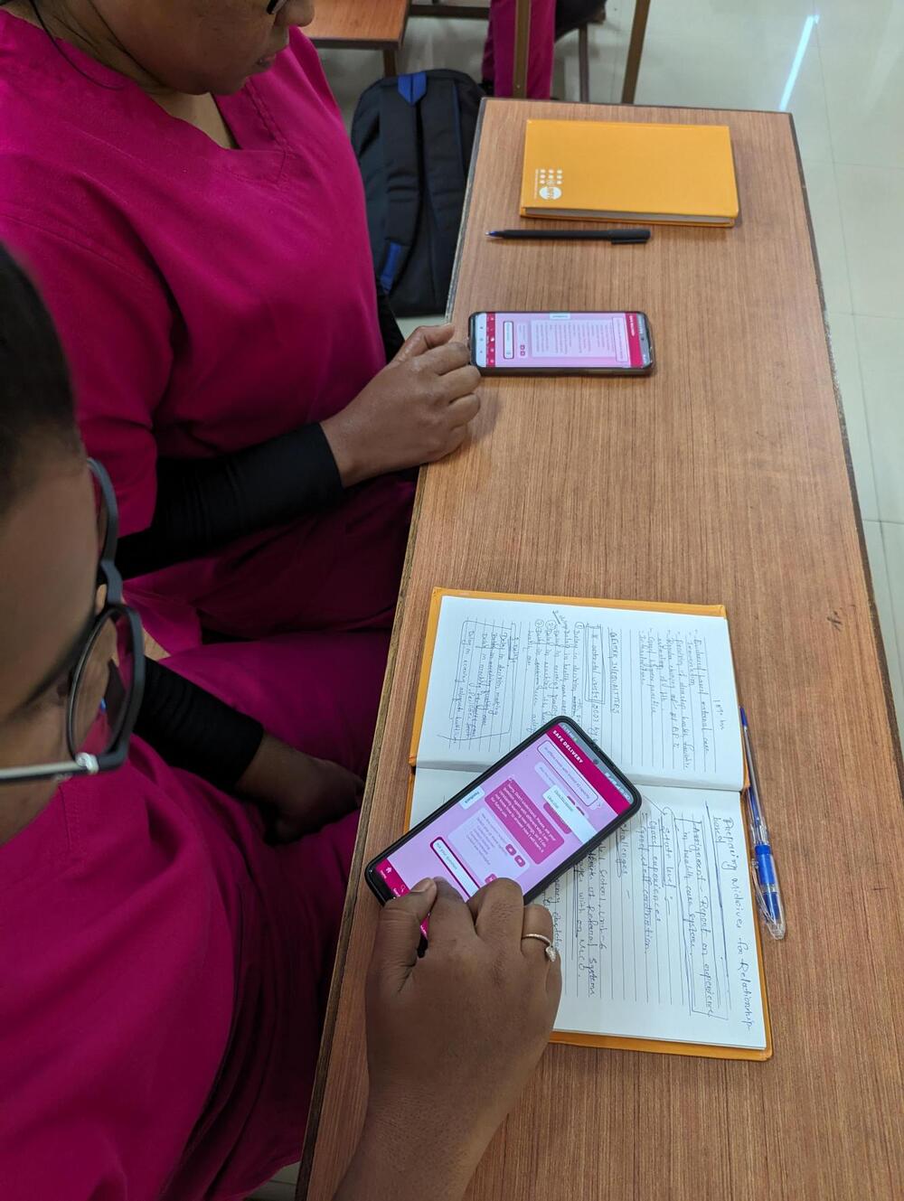 Deux femmes utilisent leur téléphone, assises à un bureau sur lequel sont posés des cahiers. 