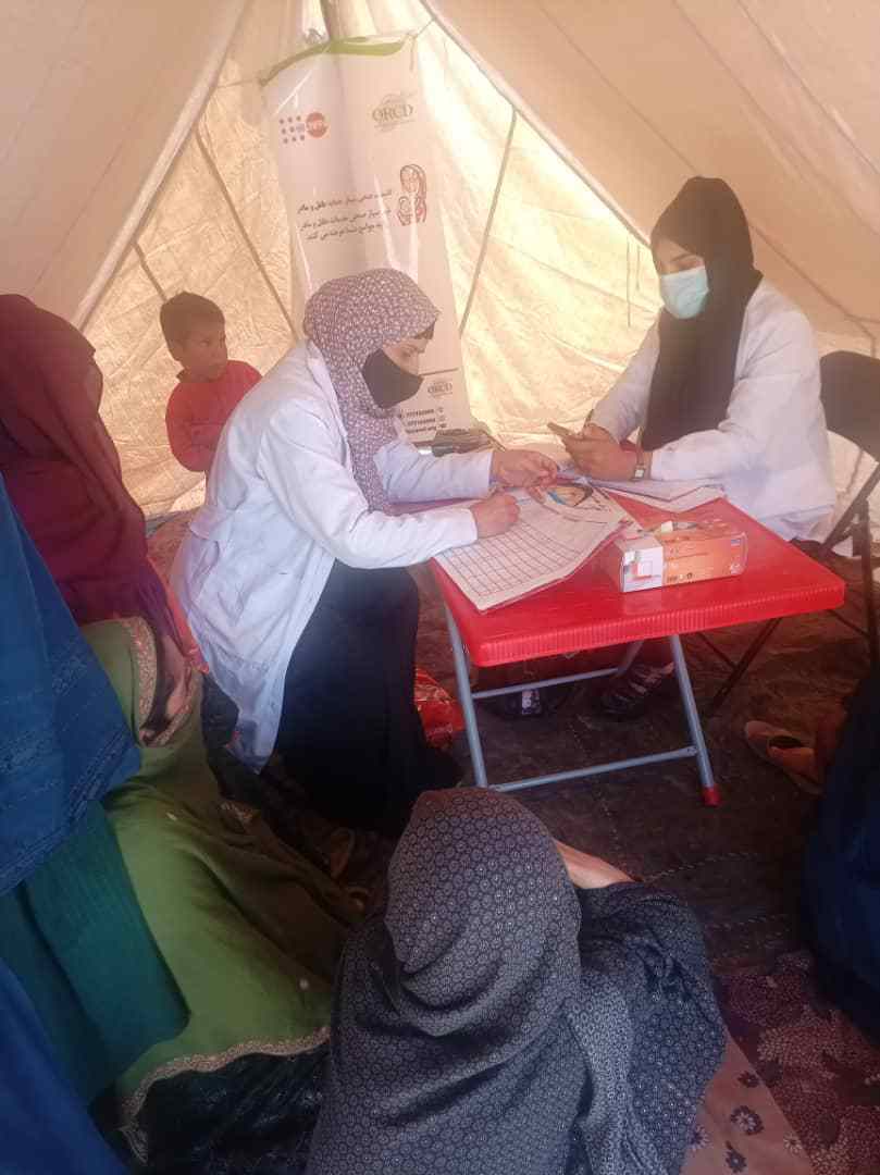 Two female health workers seated inside a tent, with two female patients and a child.