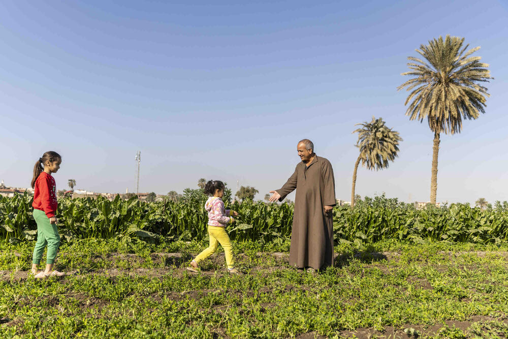 Un homme vêtu d’une tunique marron tend la main vers deux petites filles qui s’avancent vers lui. Derrière ces trois personnes, on voit deux palmiers et de la végétation verte.