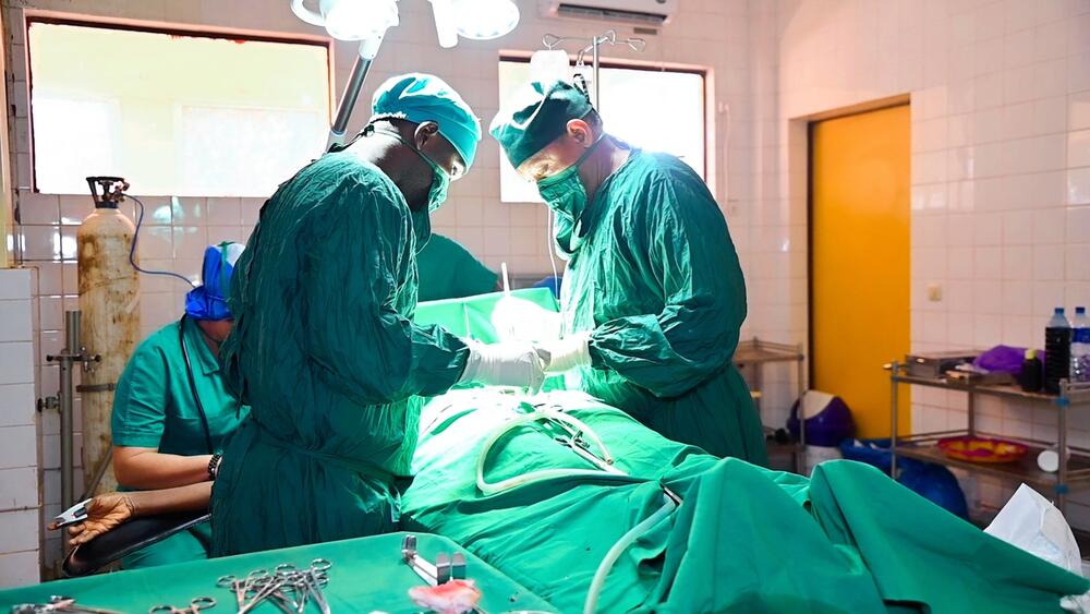 Two surgeons in scrubs operate on a patient lying on an operating table. A third health-care worker sits behind them. 