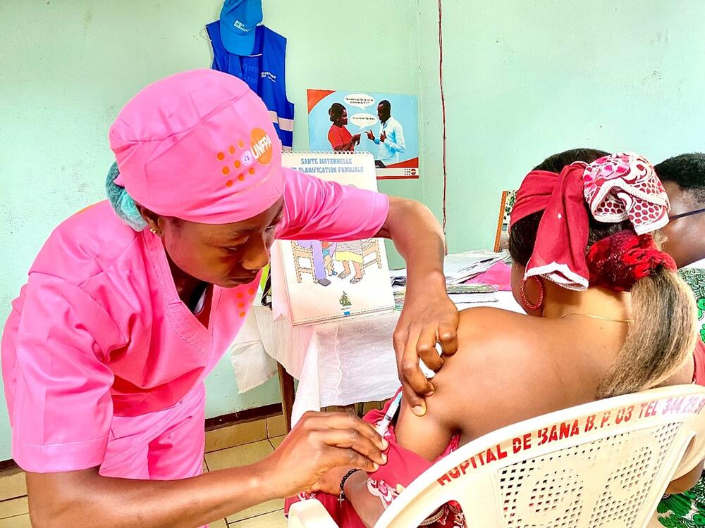 A woman in a pink uniform administers an injection into the arm of a seated woman, seen from the back