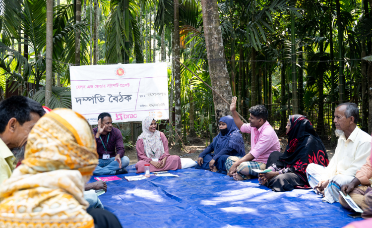  A group of men and women sit around a tarpaulin sheet, surrounded by trees.