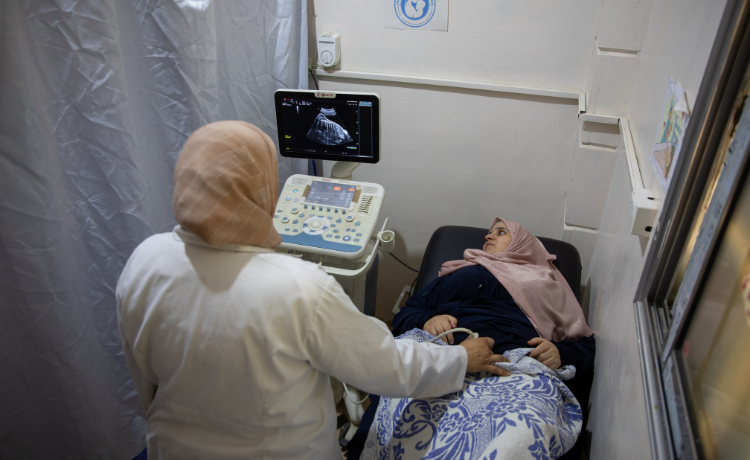 A woman lies on a hospital bed looking up at a screen showing a scan of her fetus, while a female doctor performs an ultrasound