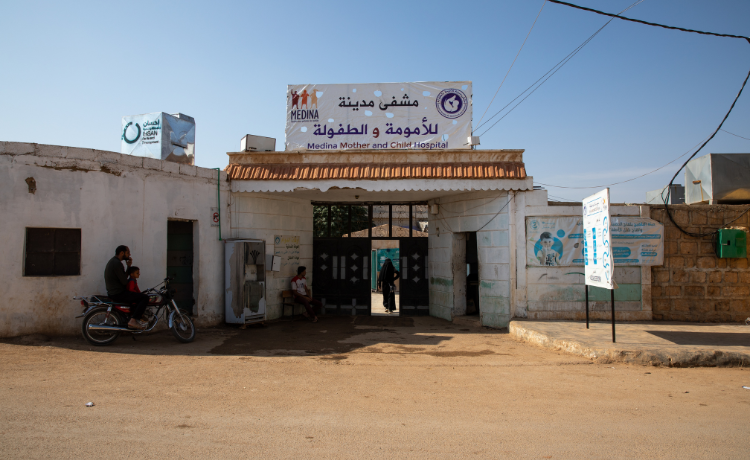 L’extérieur de l’hôpital maternel et néonatal Medina ; on voit un homme et un enfant attendre sur une moto, et une femme passant le portail d’entrée