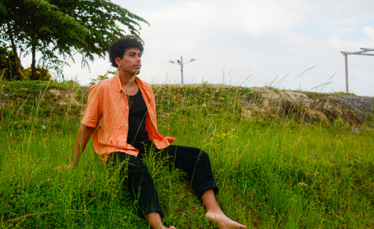 A barefoot young man in an orange shirt sits on a grassy hill