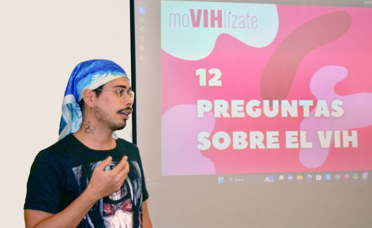 A young man with glasses stands beside a powerpoint presentation on a large projector screen