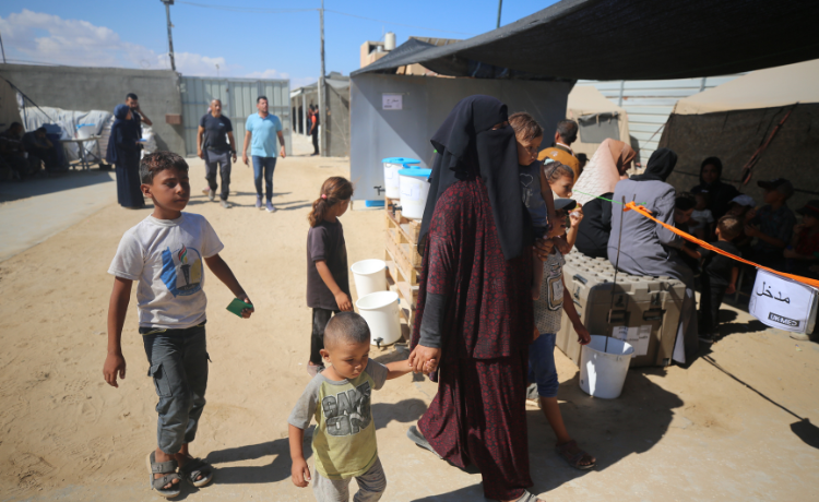 Une femme enceinte tient un petit enfant par la main, suivie par un enfant plus âgé ; elle marche sur le sable près de plusieurs tentes. On aperçoit d’autres personnes à l’arrière-plan