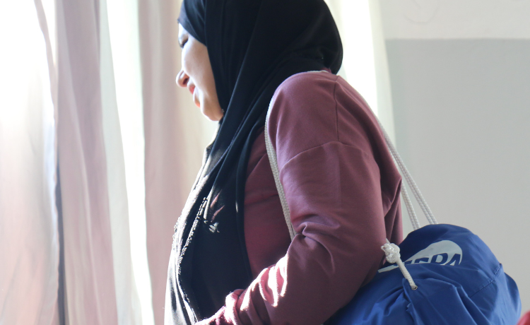 A woman wearing a dark headscarf and pink sweater carries a blue drawstring bag with a white UNFPA logo.