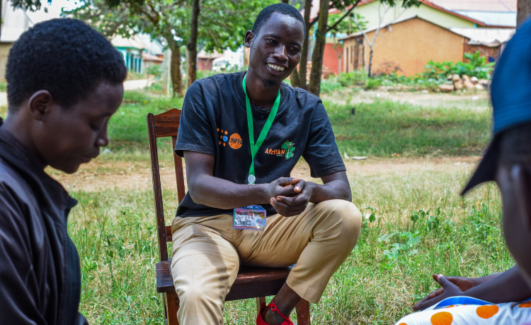 Un homme en t-shirt noir portant le logo orange de l’UNFPA est assis sur une chaise dans une cour, à l’ombre, et s’adresse à un groupe de jeunes.