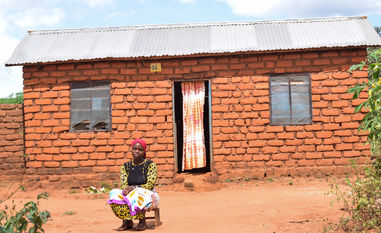  A woman wearing a red hair covering sits in front of a brick home with a corrugated metal roof.