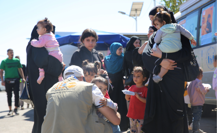 A man in a UNFPA vest hugs a child in the foreground. To his left is a woman holding a toddler