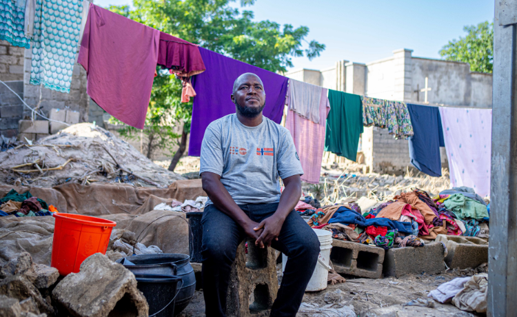 Un hombre sentado en un bloque de hormigón frente a los escombros provocados por las inundaciones y unas prendas que cuelgan de un tendedero. Lleva una camiseta gris con el logotipo del UNFPA