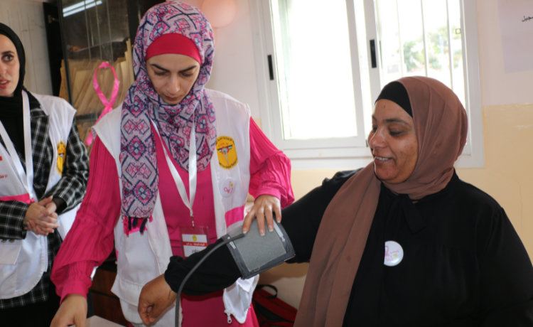 A woman in pink holds onto a blood pressure cuff, which is on the arm of a woman wearing a brown headscarf and black dress.