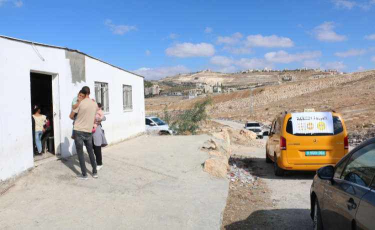 An orange van carries the UNFPA and Palestinian Medical Relief Society logos on a banner covering the back window. It is parked outside a white concrete building in a rural area.