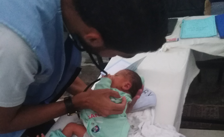  A male health worker in a blue vest leans over a newborn baby on a hospital bed, wrapped in a light green cloth