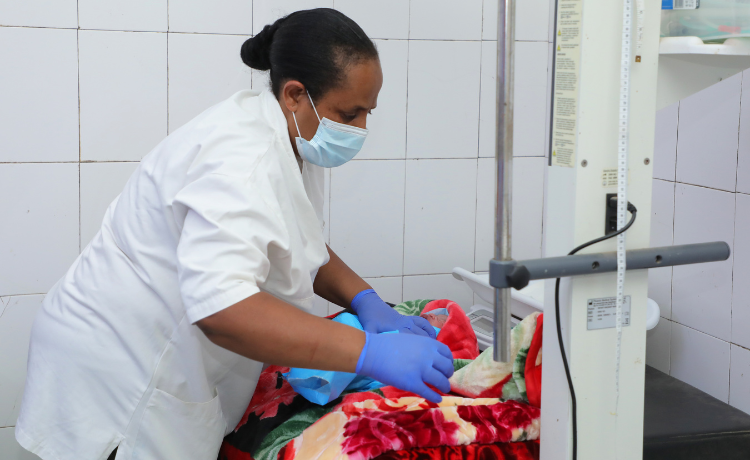 A woman in a white medical coat and blue surgical mask and gloves attends to a newborn baby wrapped in colourful blankets in a hospital ward.