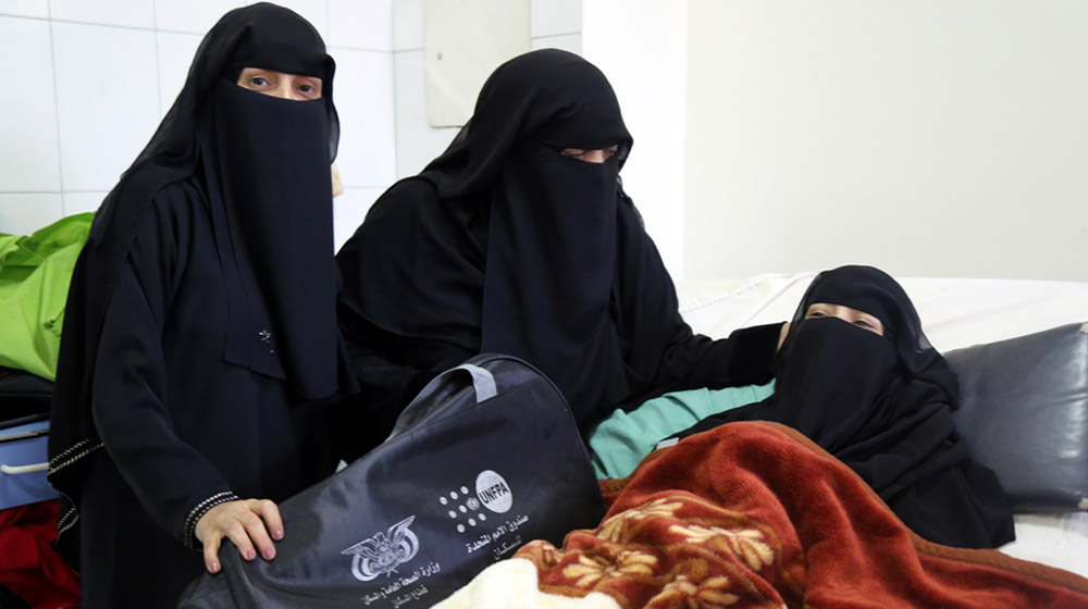 A women lays on a bed. She is attended to by two other women. All three women are in black burkas