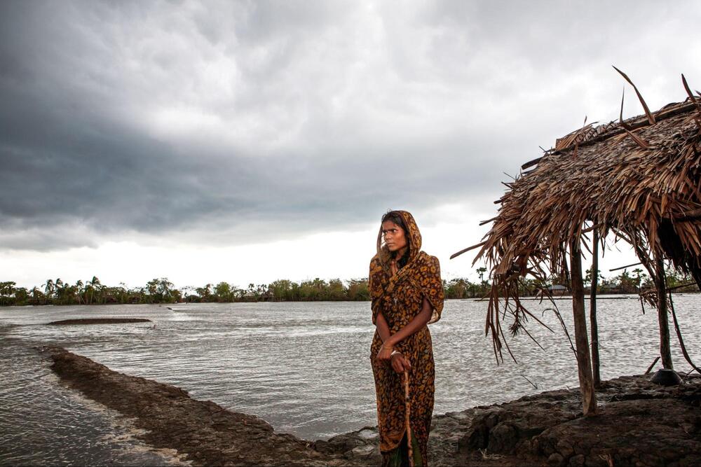 Une jeune femme est debout près d’un abri à toit de chaume et regarde par-delà des terrains inondés]